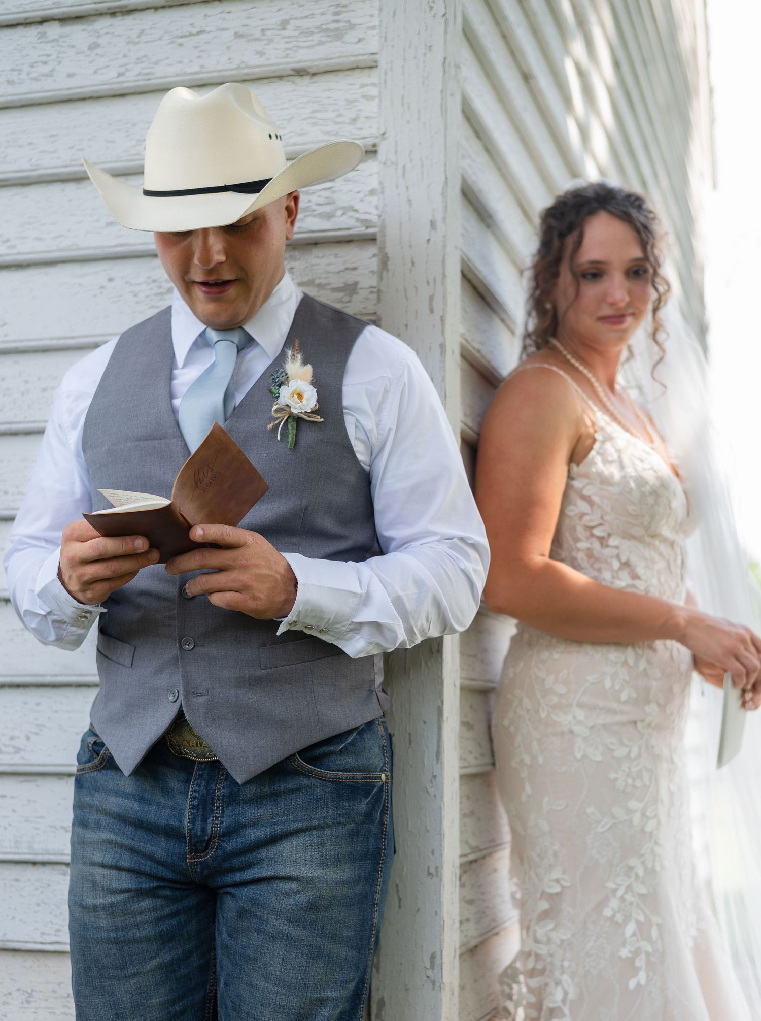 A couple in wedding attire stands together reading from books during an outdoor ceremony.