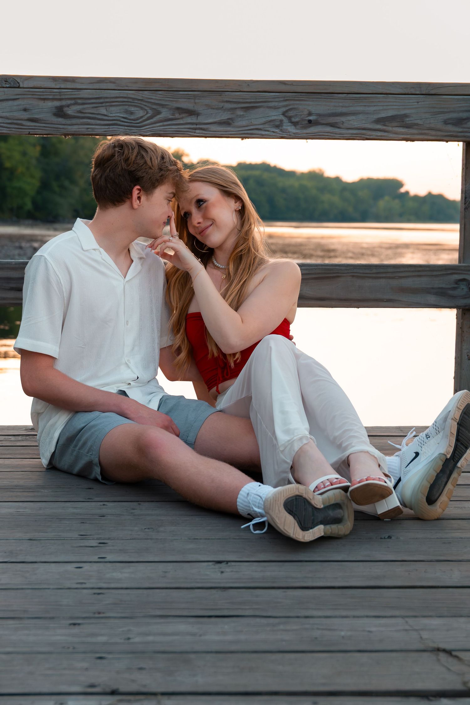 A young couple sits together on a wooden dock by a lake at sunset, sharing a romantic moment.