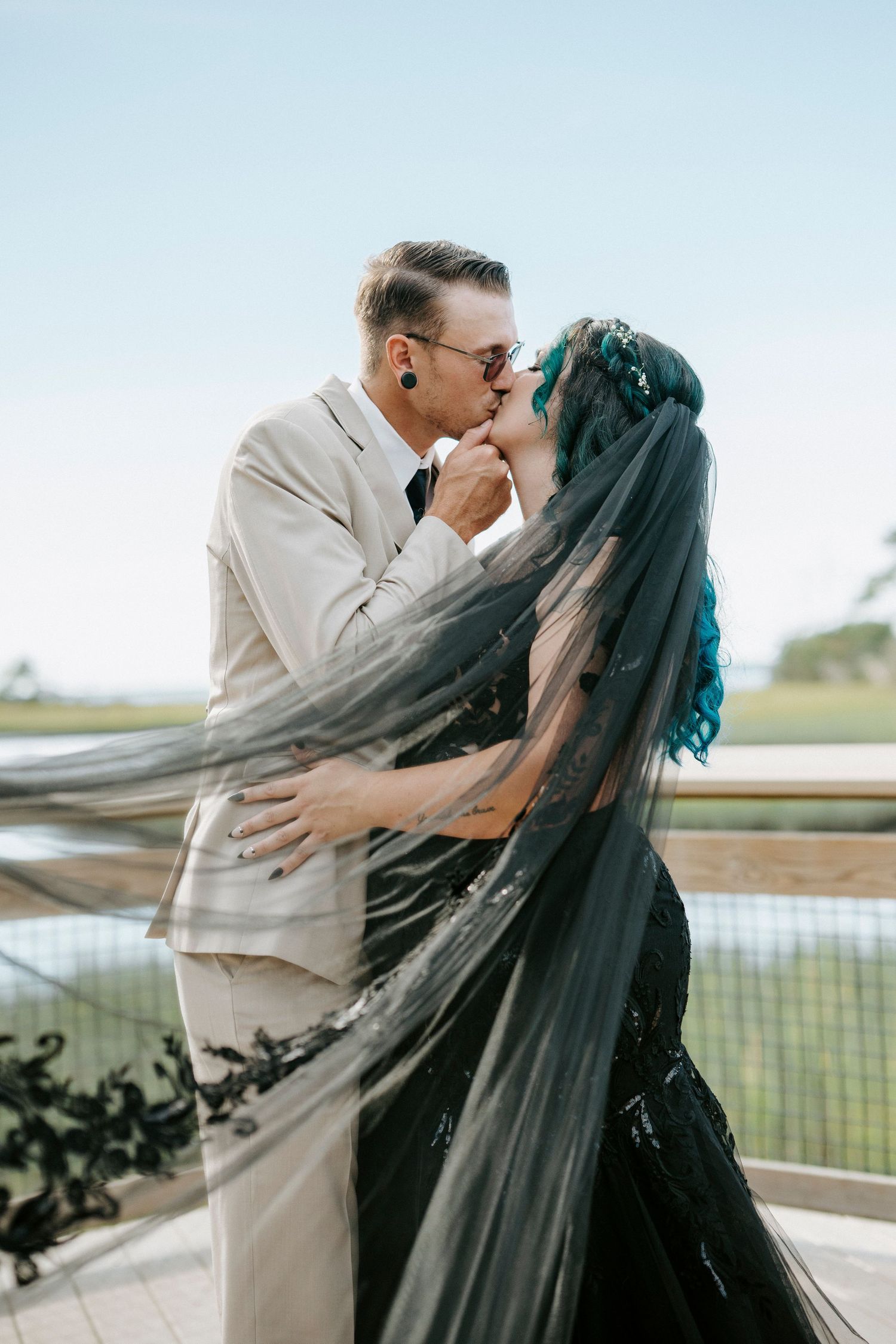 A romantic wedding couple sharing a kiss under a flowing black veil against a soft blue sky.