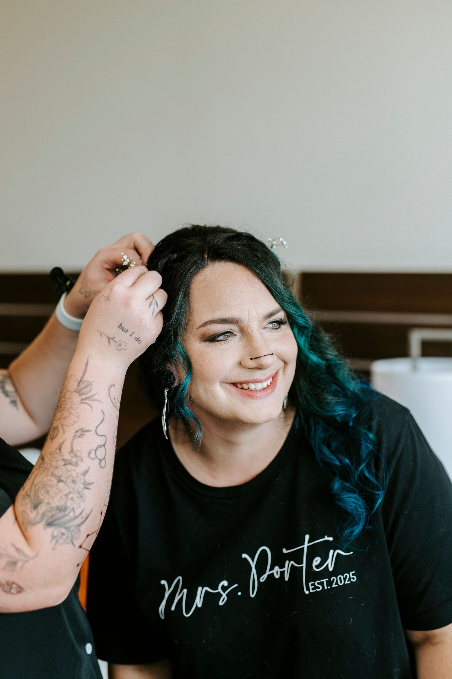 A happy individual with teal-dyed hair wearing a black Mrs. Porter t-shirt and showing off arm tattoos.