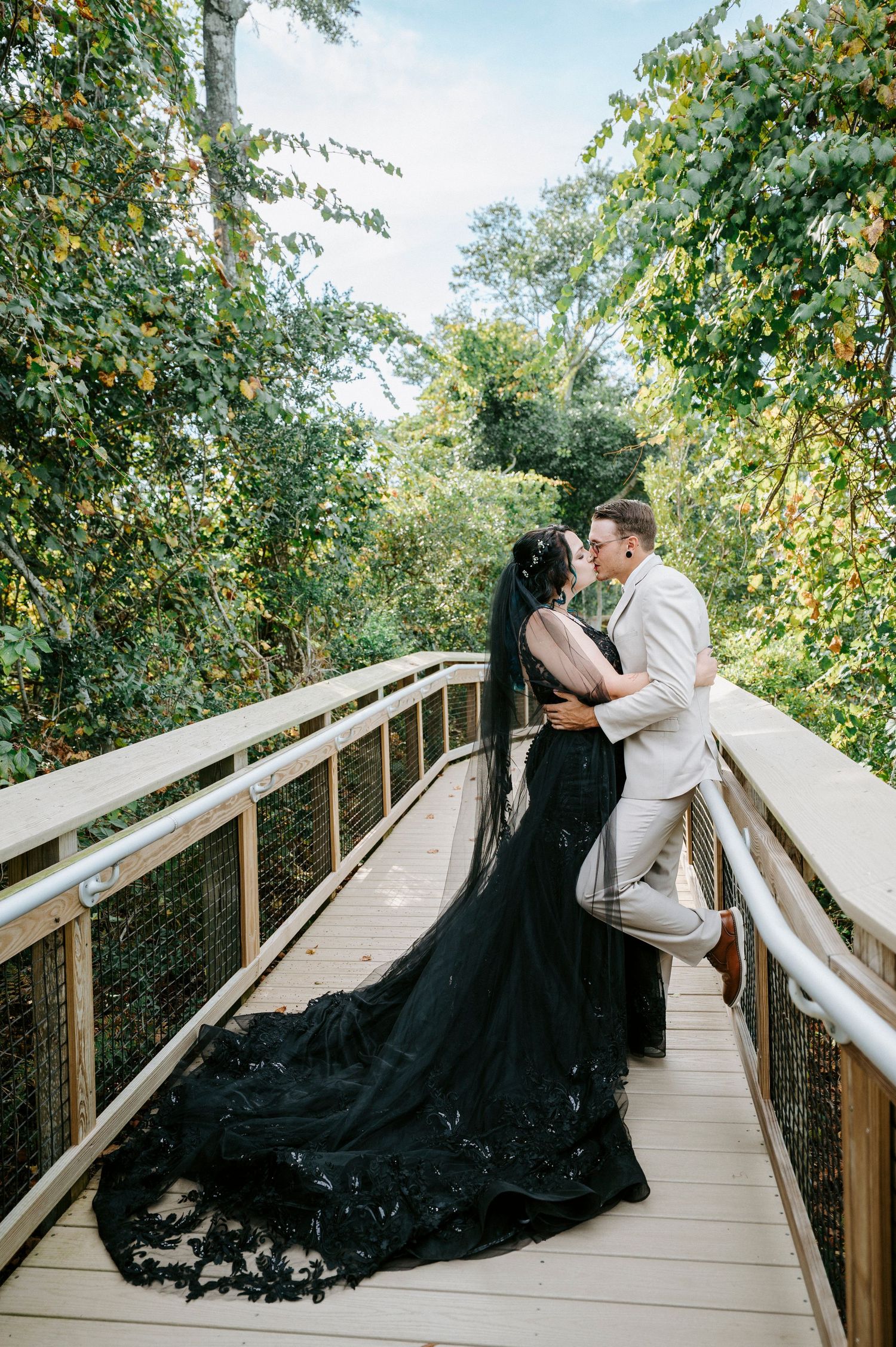 A couple in contrasting beige and black outfits pose dramatically on a wooden boardwalk surrounded by greenery.