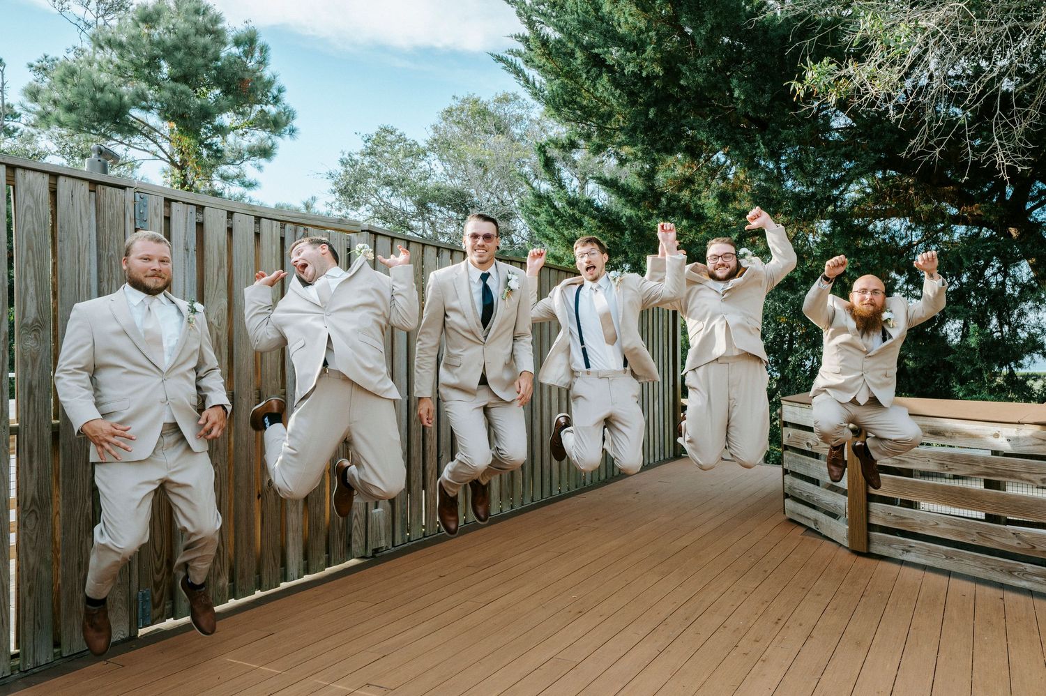 Wedding party members in light gray suits and brown shoes pose together on wooden steps outdoors in nature.