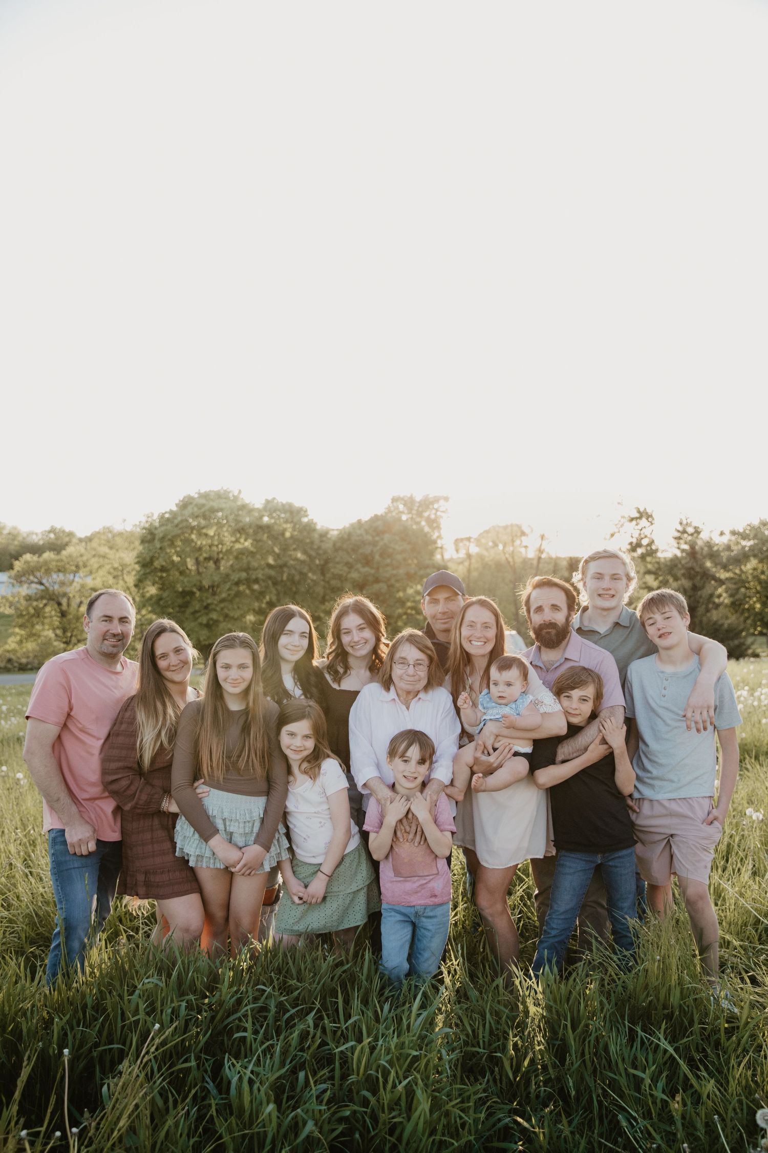 Extended family portrait in a sunny field at golden hour with trees in background.