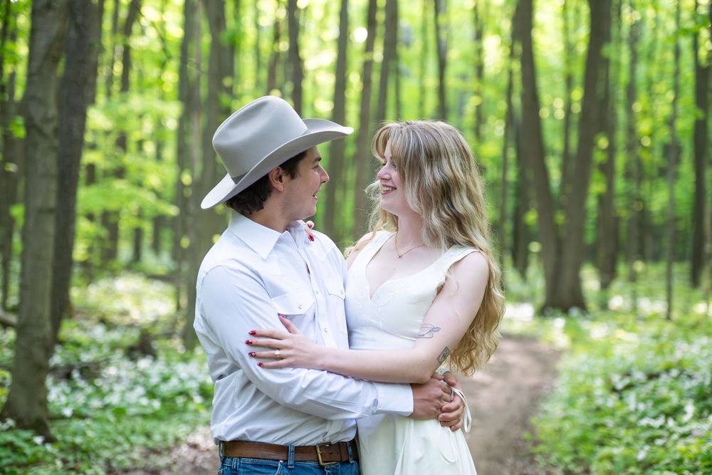 A romantic couple embraces on a forest path surrounded by blooming spring wildflowers and lush green trees.