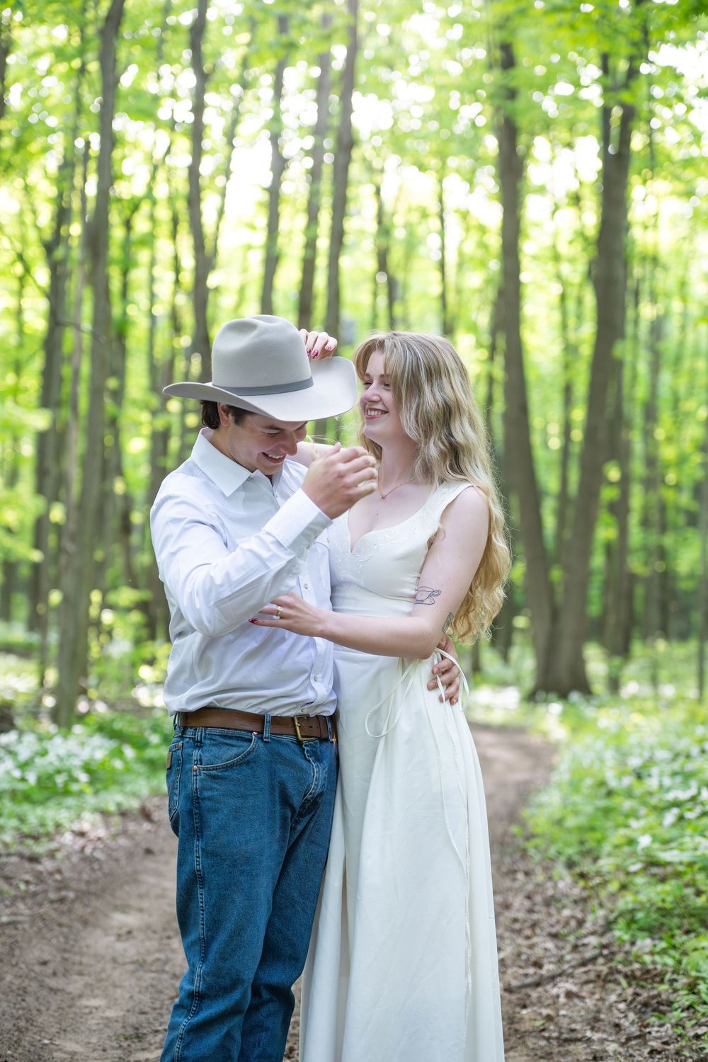 Dancing couple embraces in lush green forest setting wearing white dress and cowboy hat with jeans.