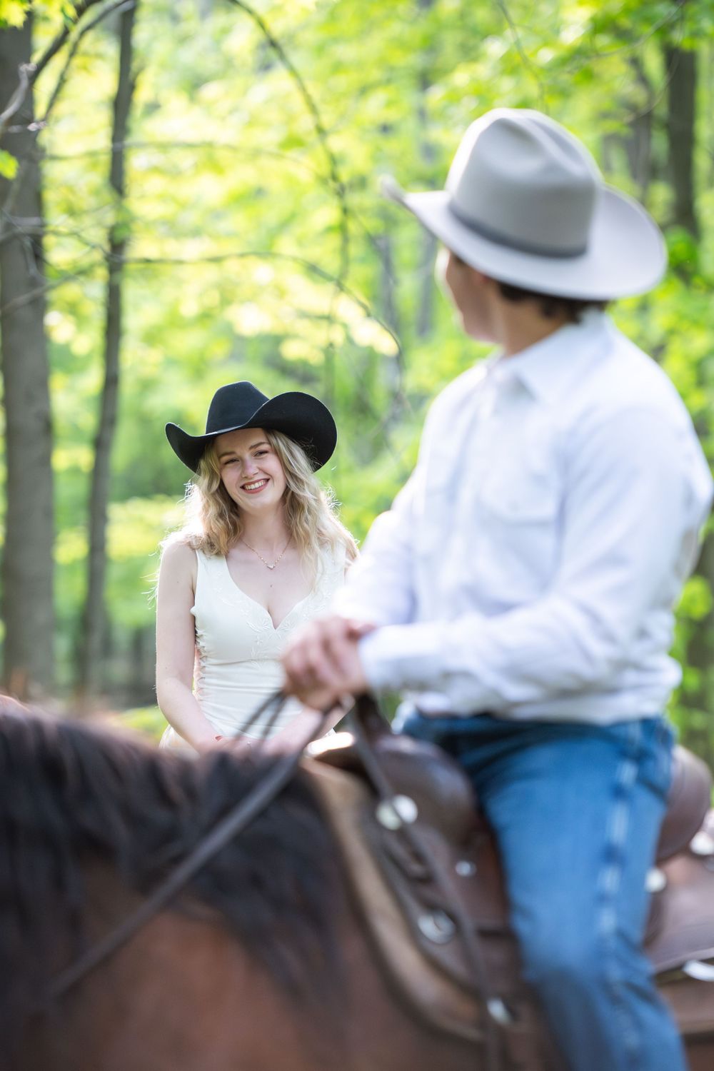 Two people in cowboy hats enjoy horseback riding through a bright green forest on a sunny day.