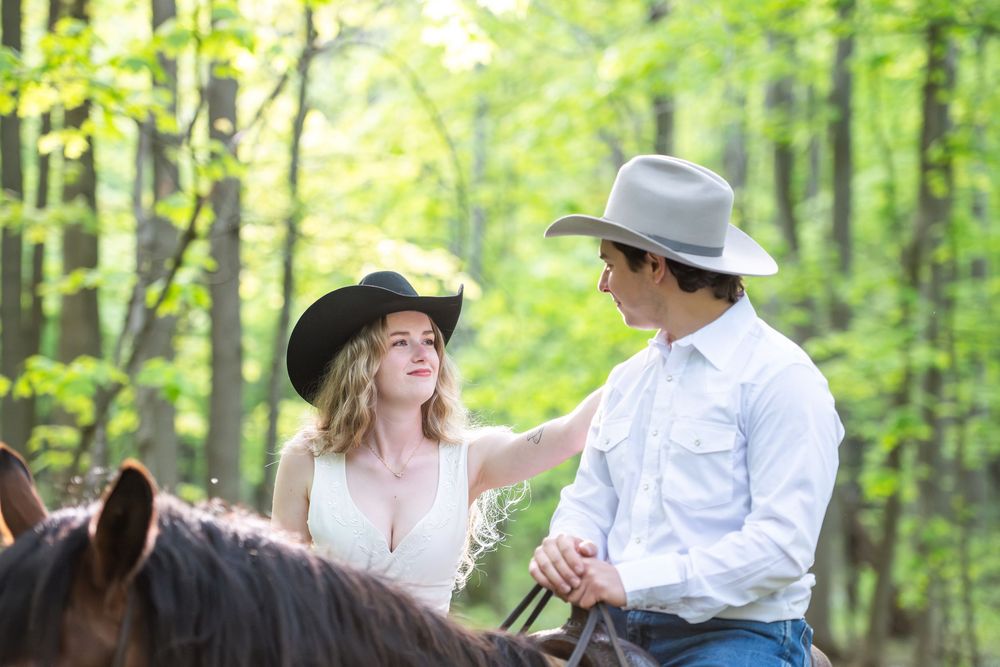 Couple riding horses together in a lush green forest while wearing cowboy hats and western attire.