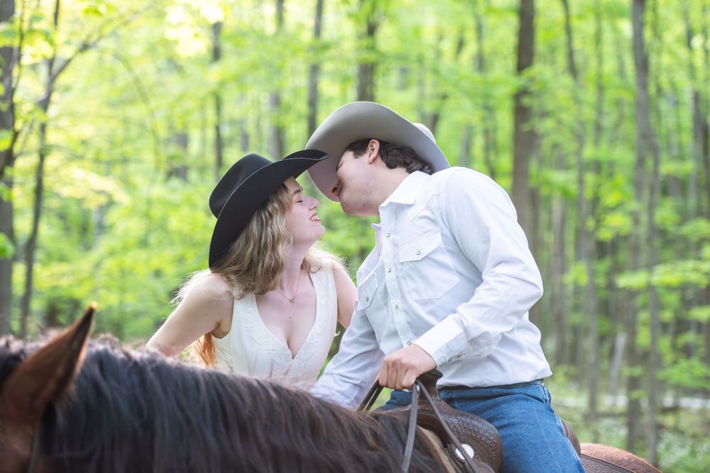 Two people ride brown horses along a wooded trail surrounded by lush spring foliage.