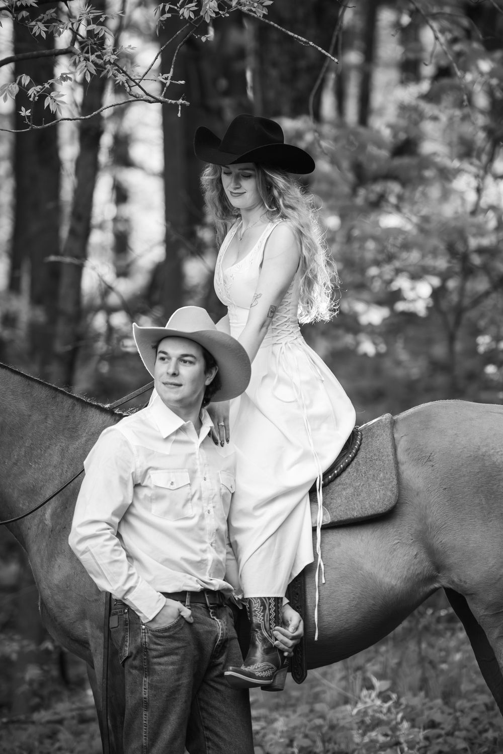 Two people in western attire pose with a horse in a black and white forest scene.