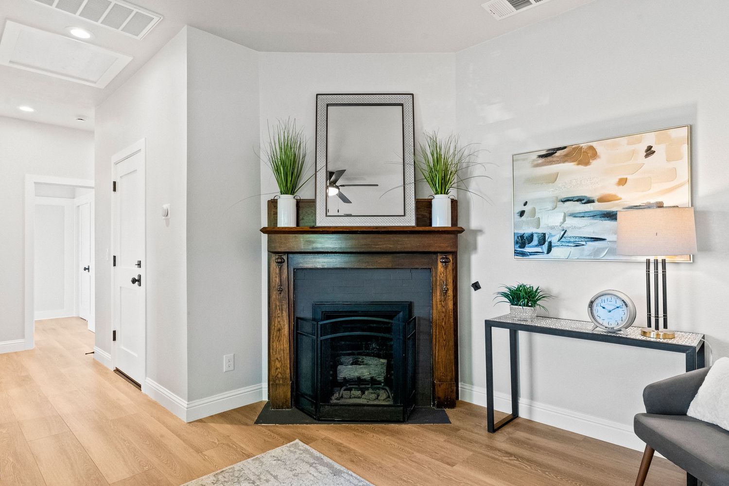 Modern living room interior with fireplace, wood mantel, abstract art, and potted plants on light hardwood floors.