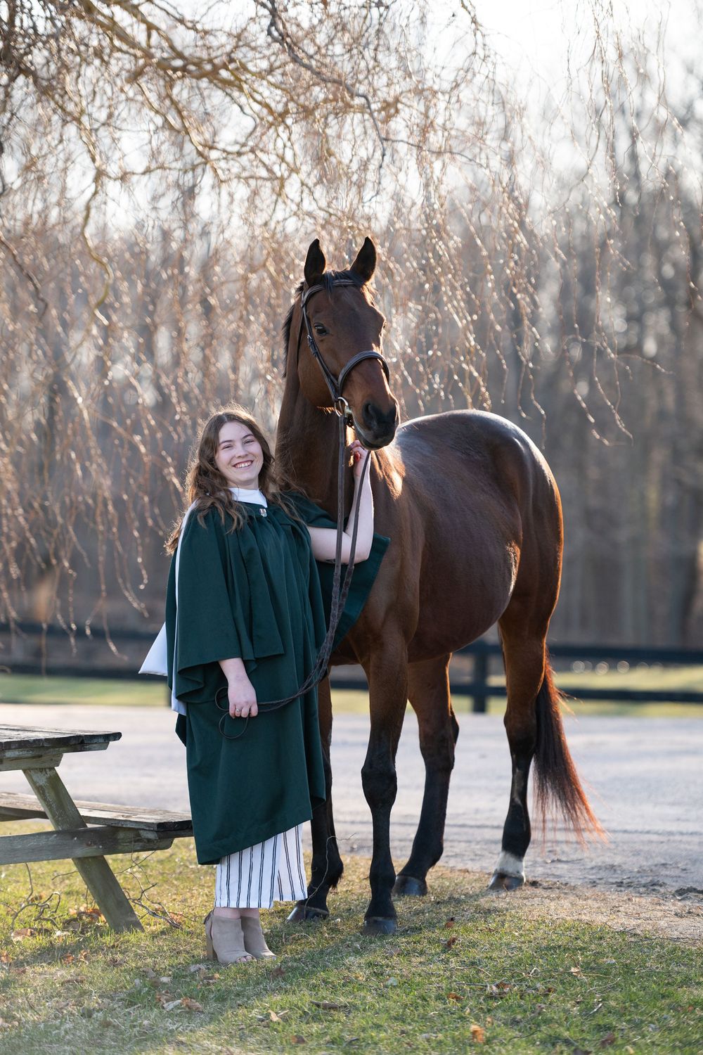 A brown horse stands with its handler wearing a green coat in a winter landscape with bare trees.