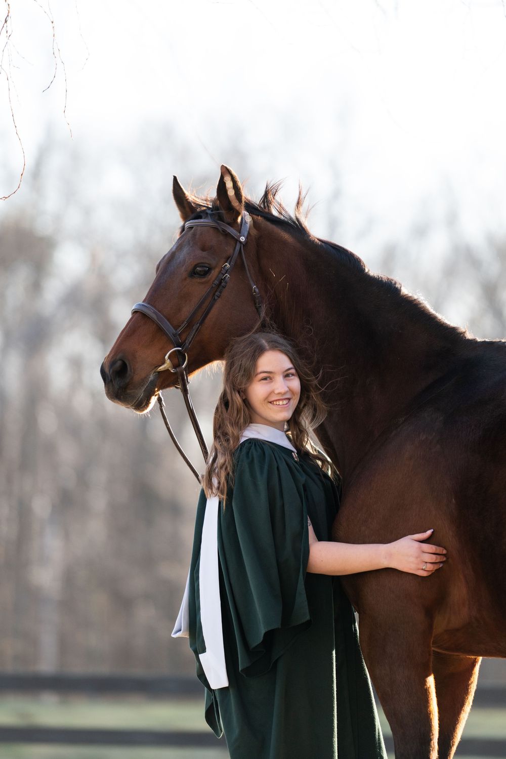 Graduate in green gown poses with a beautiful bay horse in an outdoor setting during a graduation photoshoot.