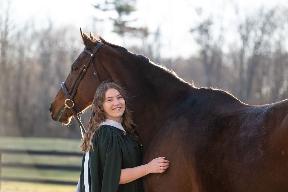 Graduate in dark green academic regalia stands beside a majestic brown horse in a winter landscape.