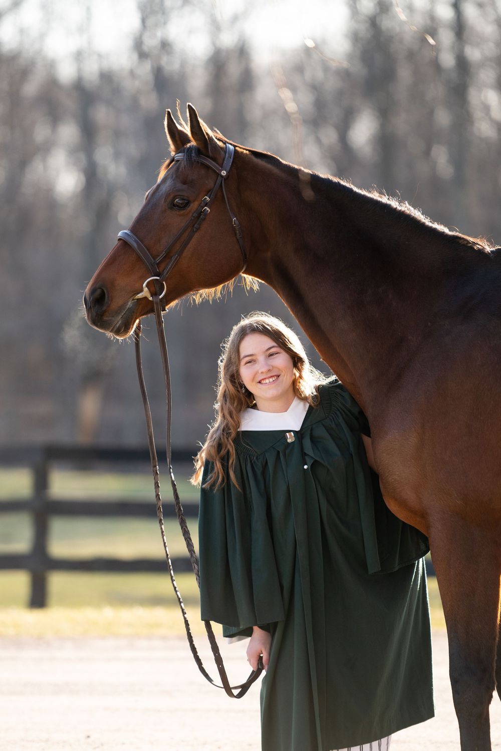 A graceful brown horse stands beside someone wearing a dark green graduation gown in a pastoral setting.