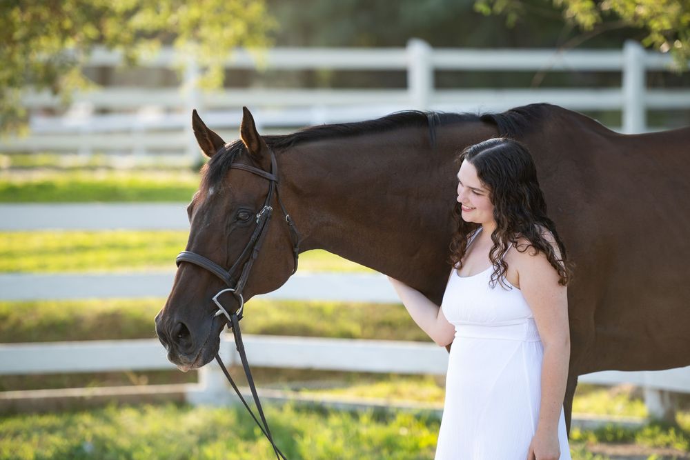 A person in a white dress poses beside a dark brown horse in a scenic outdoor setting with sunlit trees.