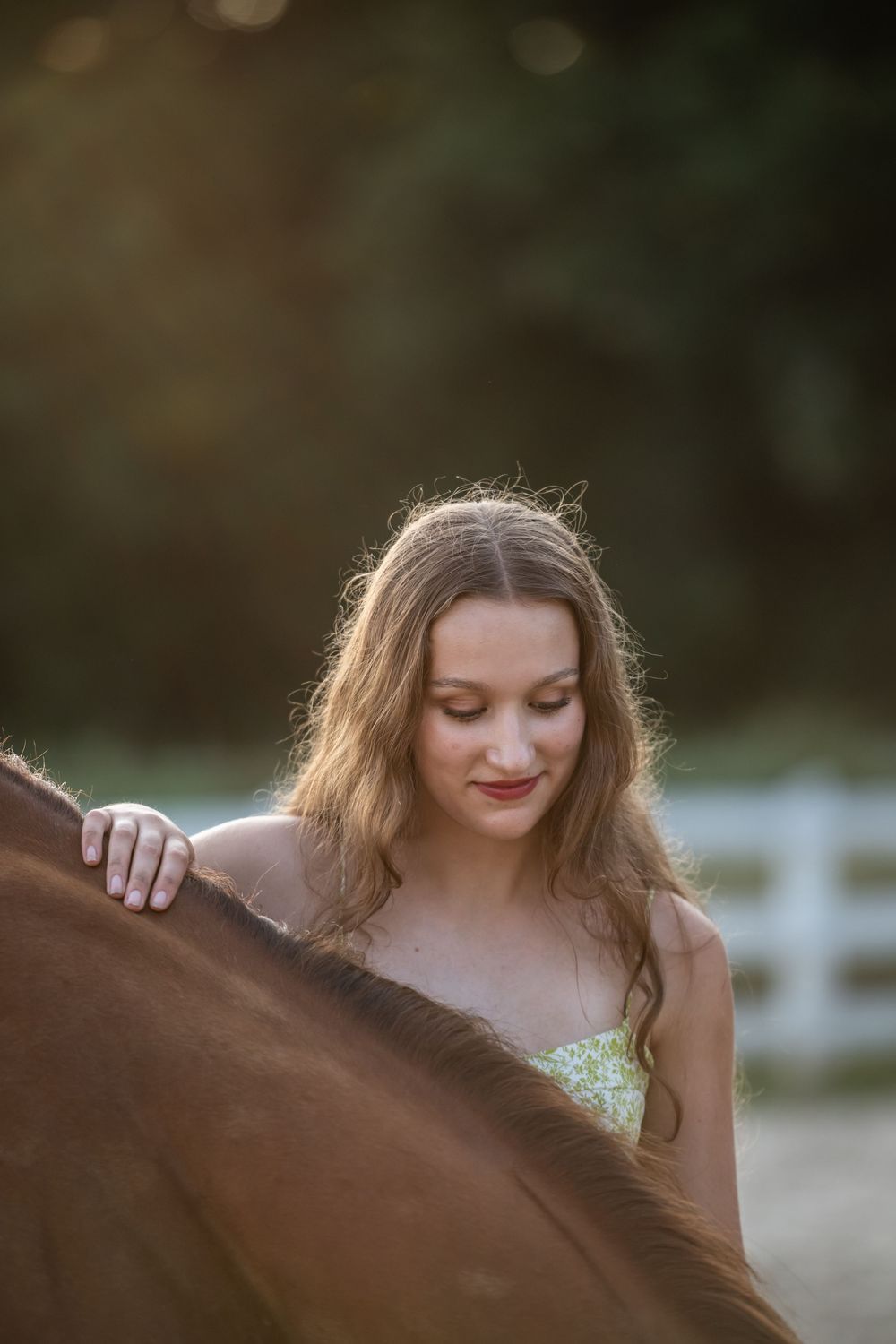 Woman in floral dress poses outdoors with a brown horse during golden hour lighting.