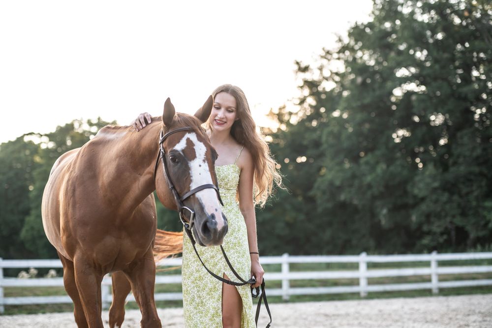 Woman walking and posing with brown horse along white fence on sunny summer evening.