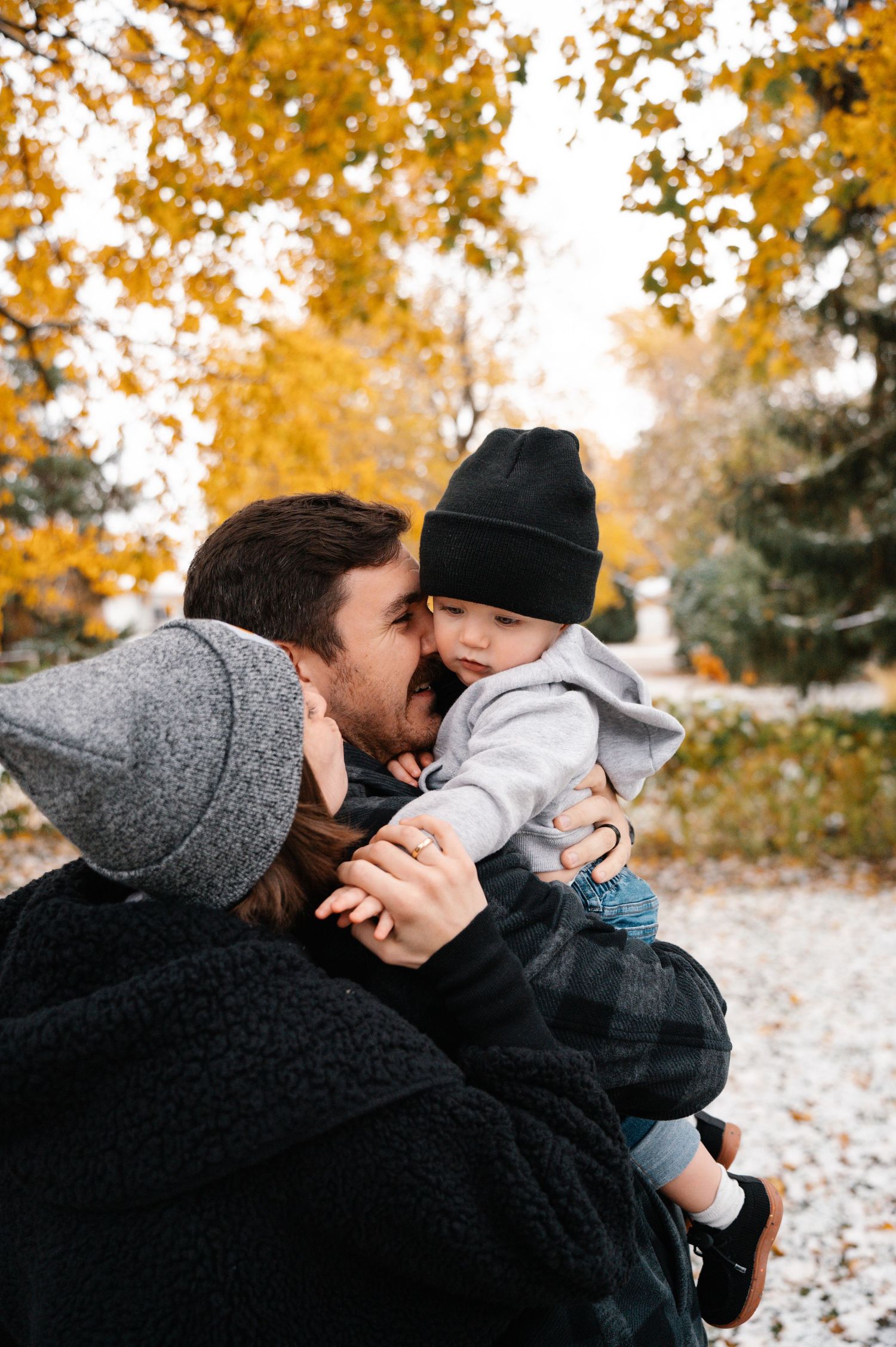 Two parents lovingly kiss their baby boy wearing a black beanie hat outdoors in fall.