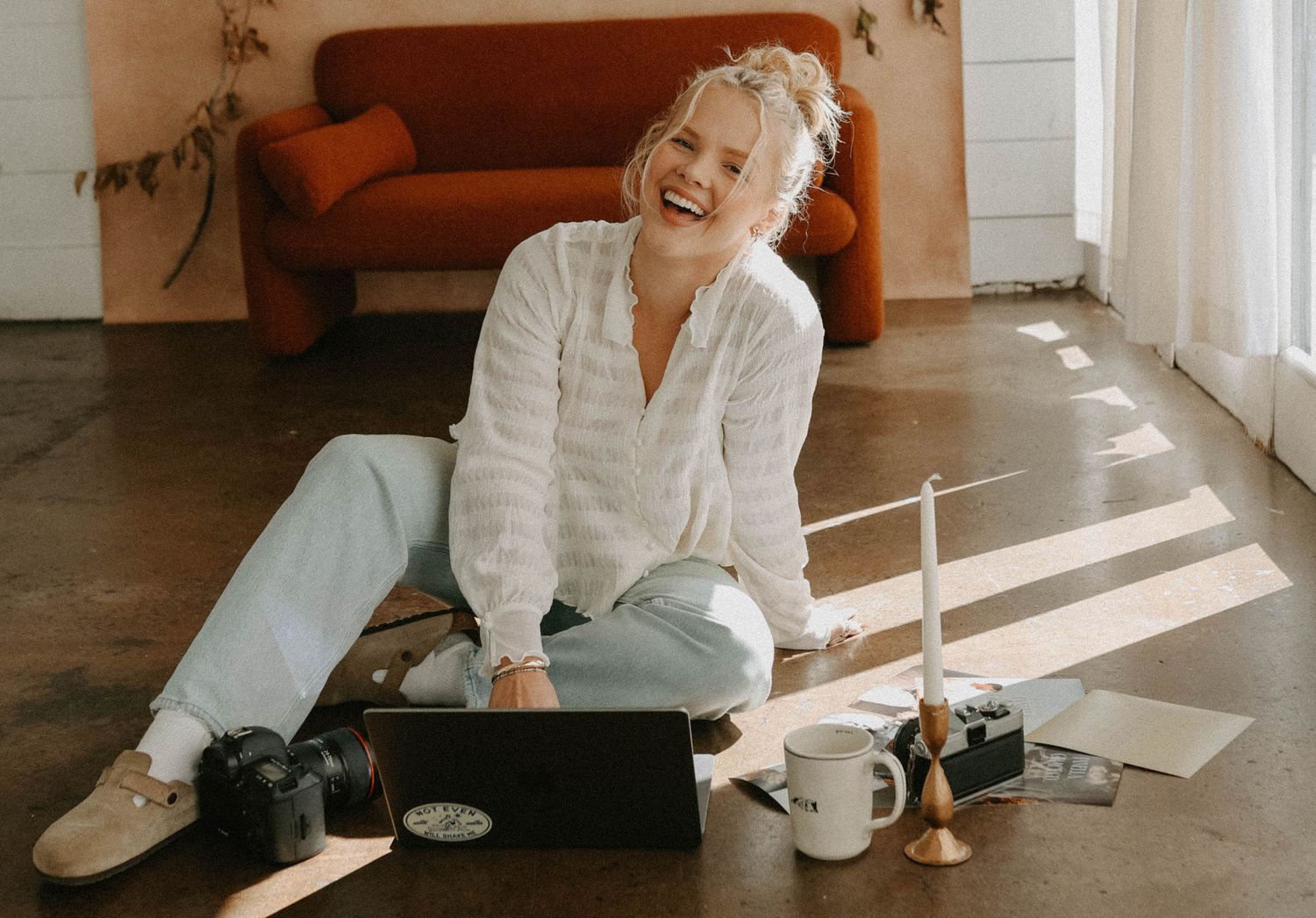 A person in a white shirt sits casually on the floor with a laptop and coffee mug in a sunlit room with orange couch.