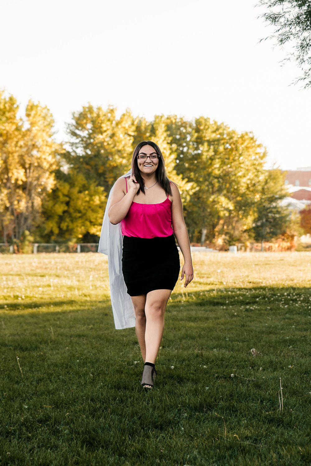 Senior with her graduation gown draped over her shoulder walks toward the camera smiling