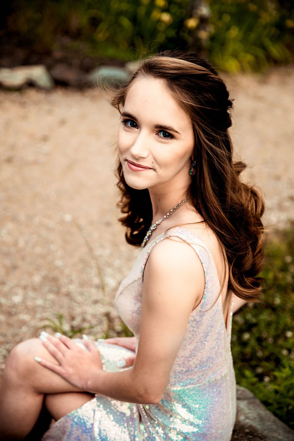 Young woman in silver Prom dress reflecting the colors of the park around her looks into the camera and smiles