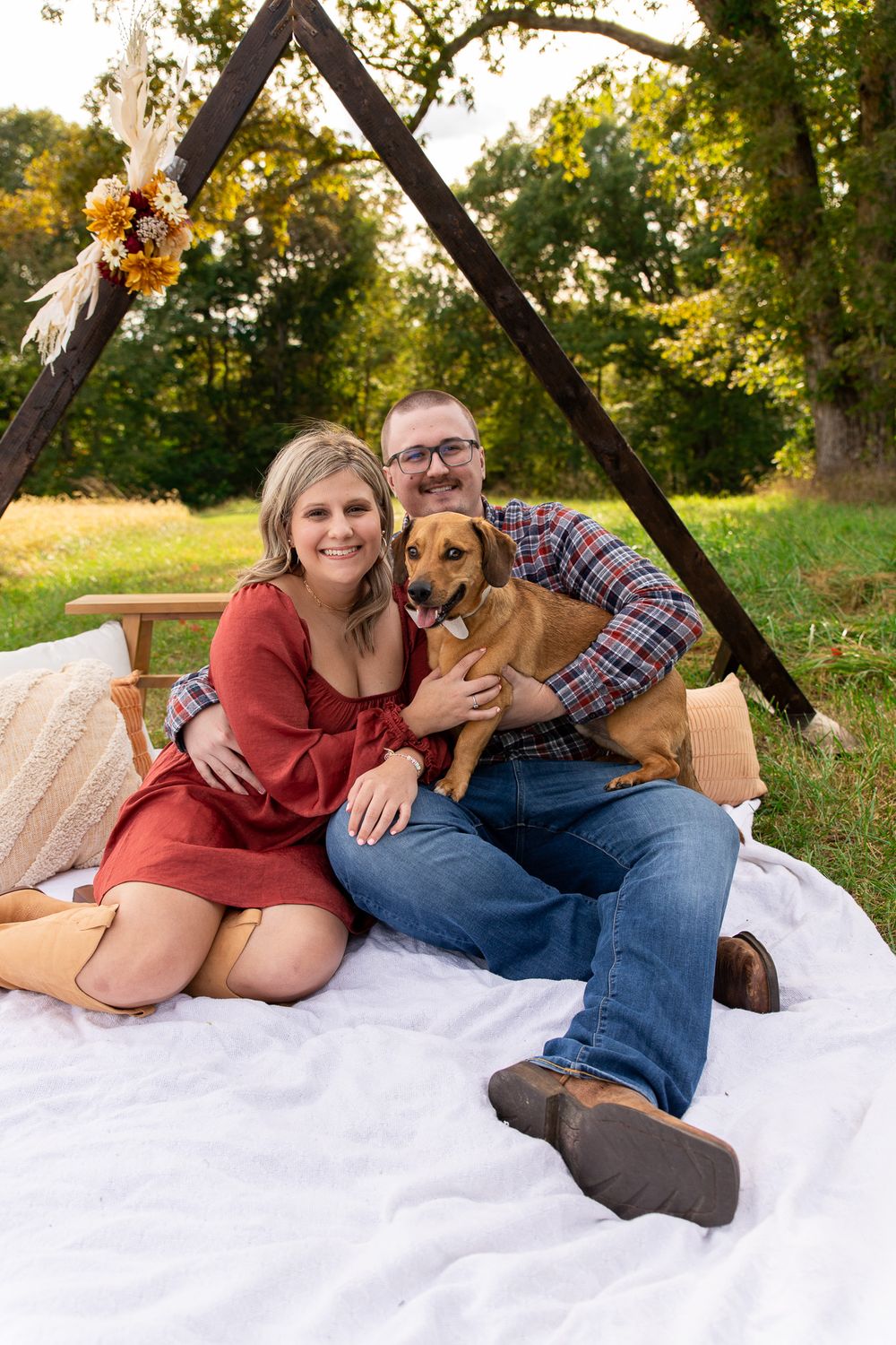 Couple relaxing on white blanket with their brown dog in sunny outdoor setting.