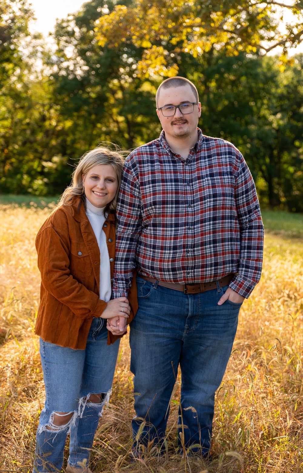 Couple poses together in a golden autumn field during sunset wearing casual plaid and denim outfits.