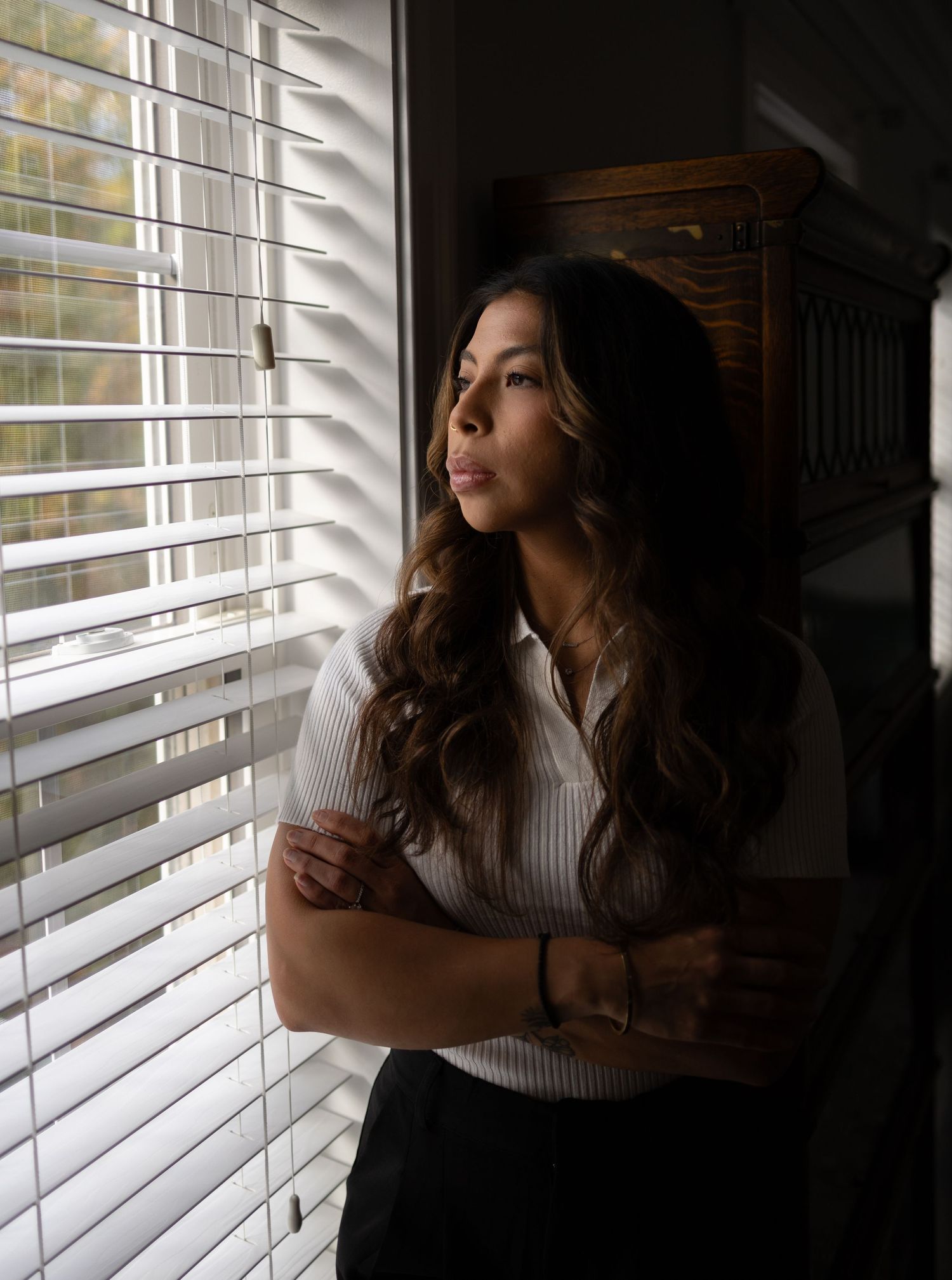 Young woman with long wavy hair gazes thoughtfully out window through venetian blinds in dramatic lighting.