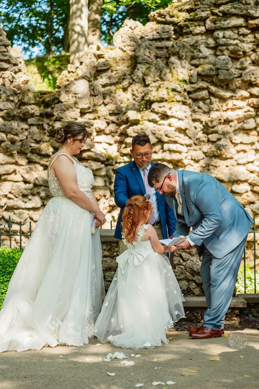 Wedding ceremony with bride, groom, officiant, and flower girl exchanging rings outdoors near a stone wall.