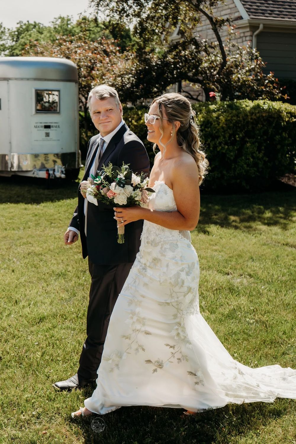 Bride in floral wedding gown and groom in black tuxedo pose together on green lawn holding bouquet.