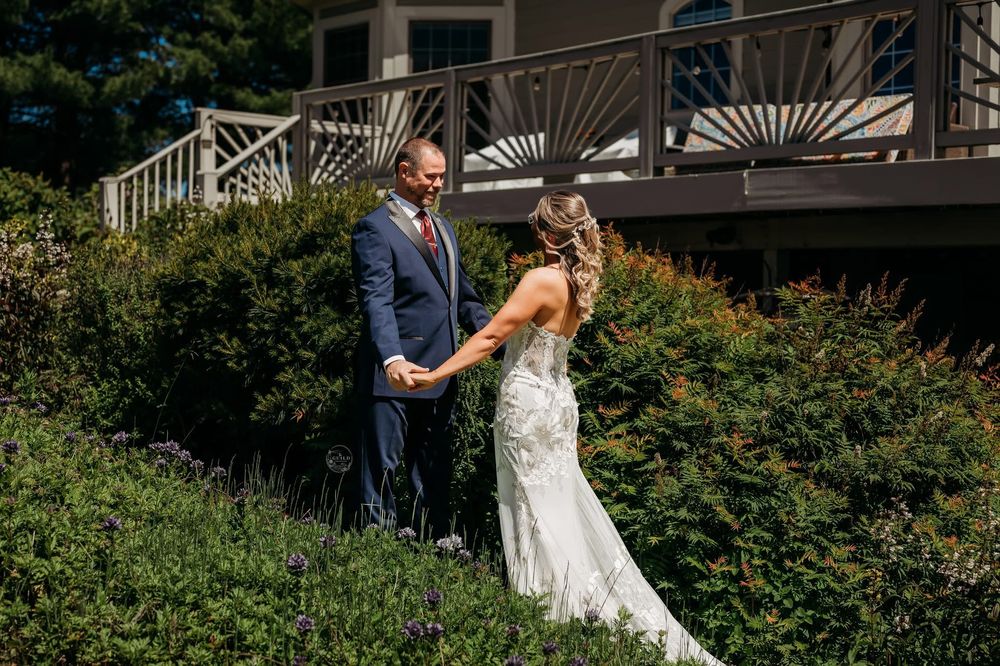 Bride and groom share a tender first look moment outdoors surrounded by lush green bushes near a wooden deck.