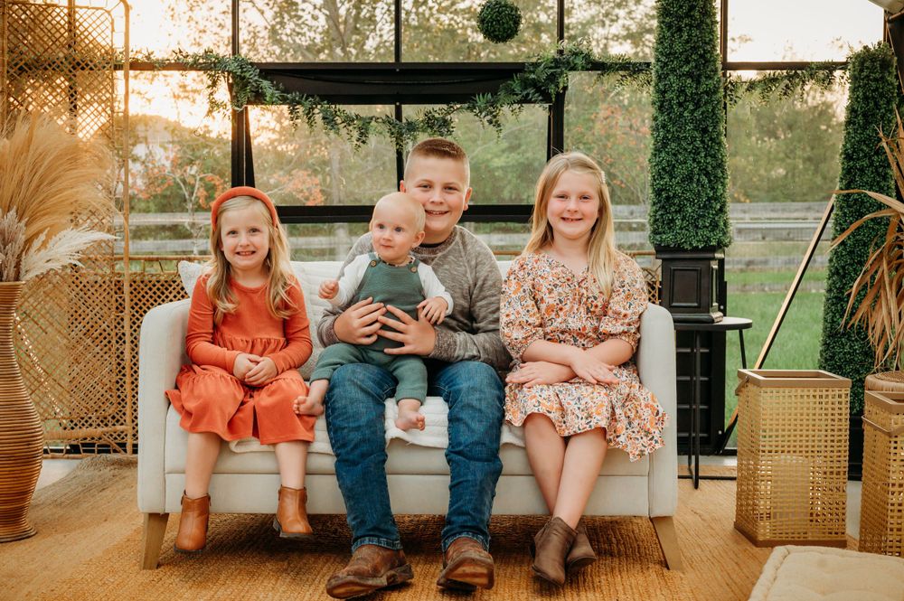 Four siblings including baby sitting together on white sofa inside sunlit greenhouse boho setting.