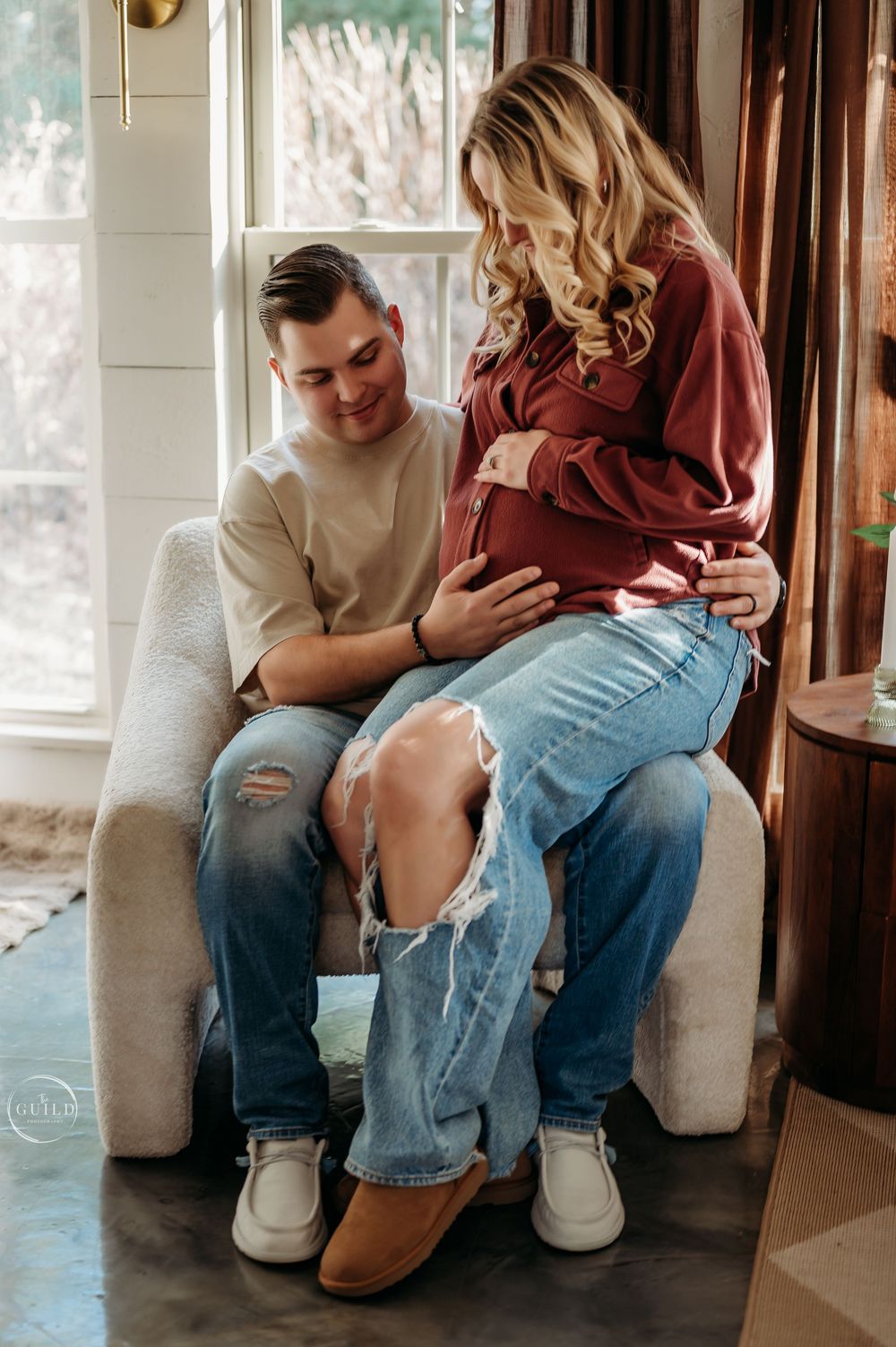 Couple sharing a tender maternity moment, sitting together in a chair, both cradling a baby bump, wearing casual jeans and cozy tops.