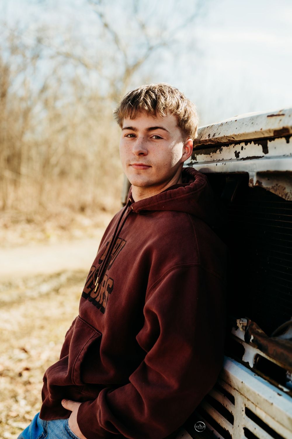 A young man in a maroon hoodie leans against a vintage vehicle outdoors in a natural setting.