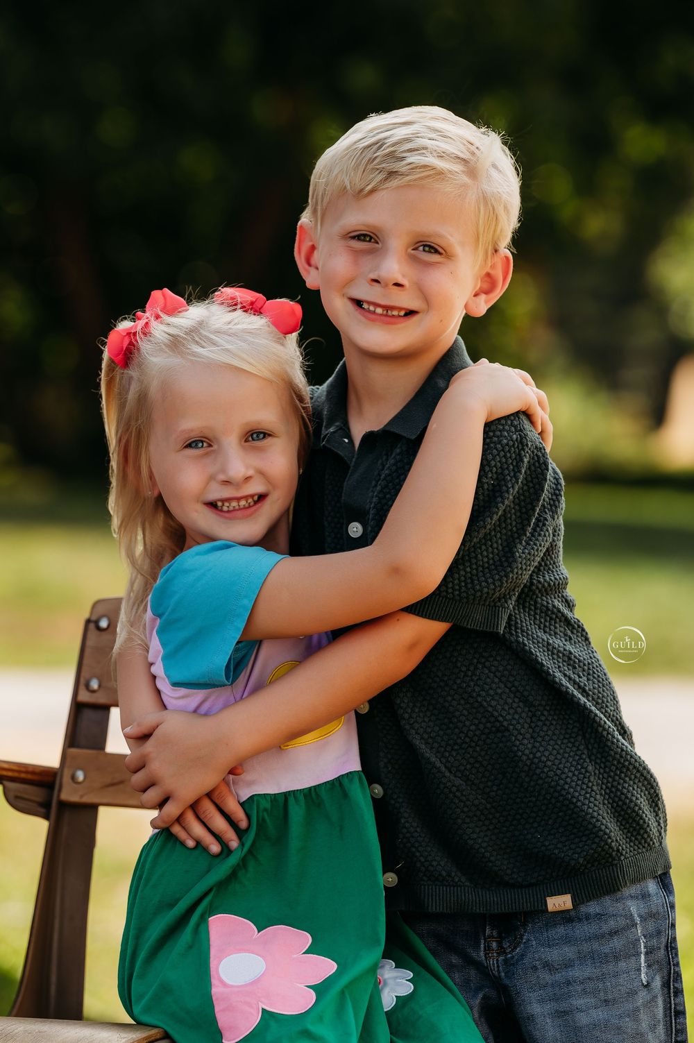 Two blonde siblings hugging and smiling outdoors, girl in green floral dress with red bows, boy in dark polo shirt.