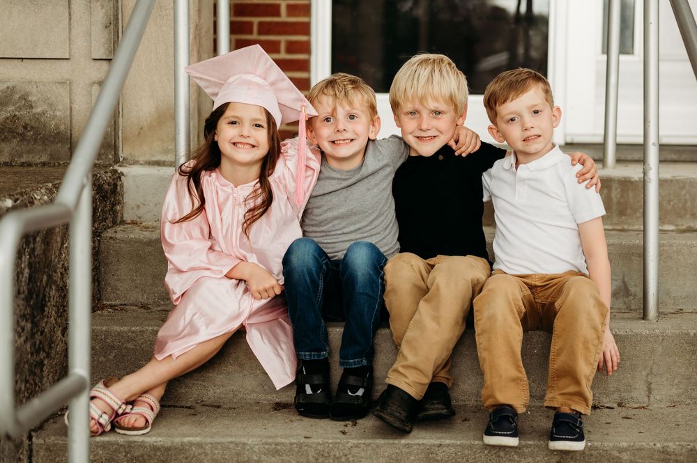Four young children smile on steps, one girl in pink graduation cap and gown with three boys in casual attire.