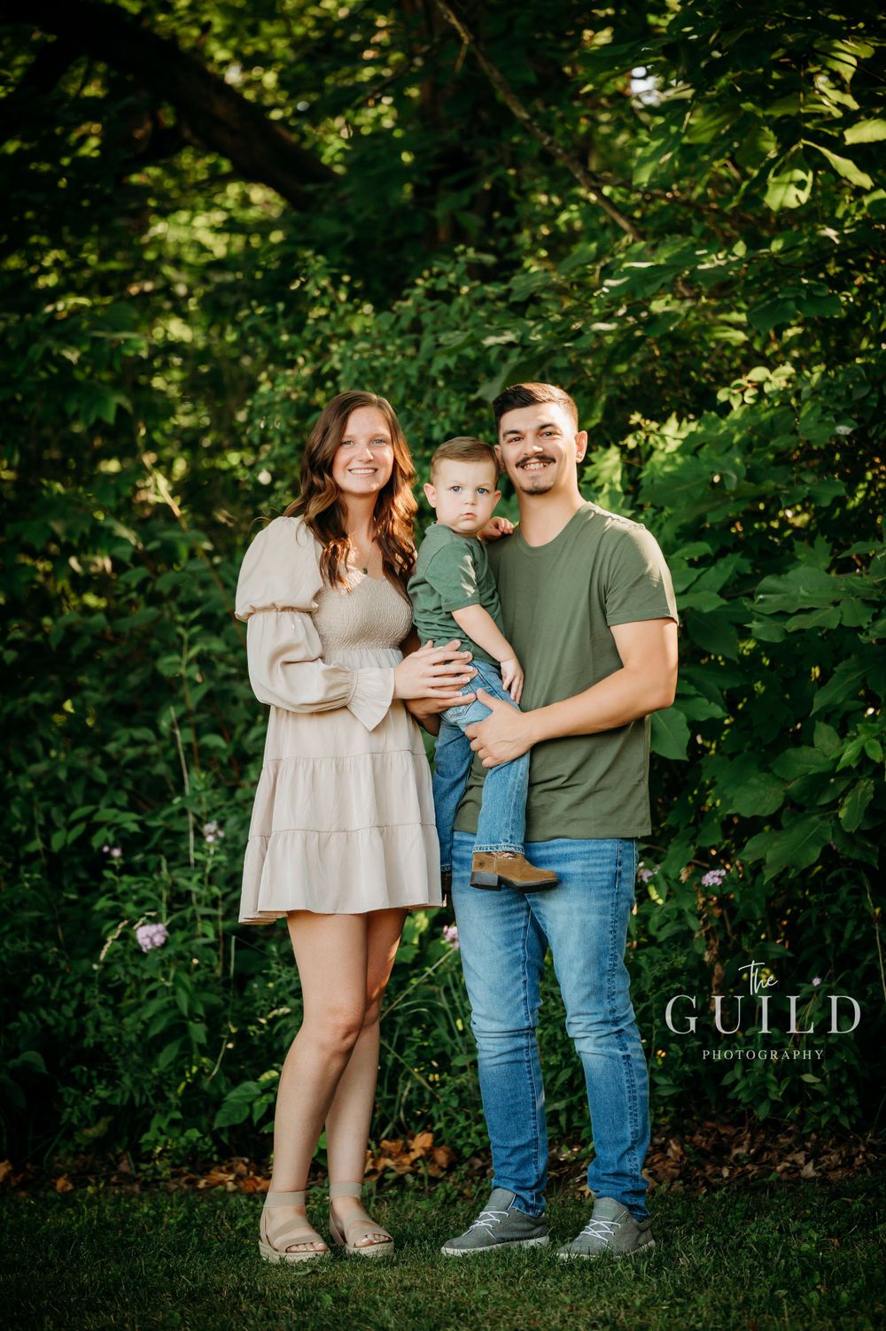 Young couple holding toddler son while posing outdoors in lush green garden for family portrait.