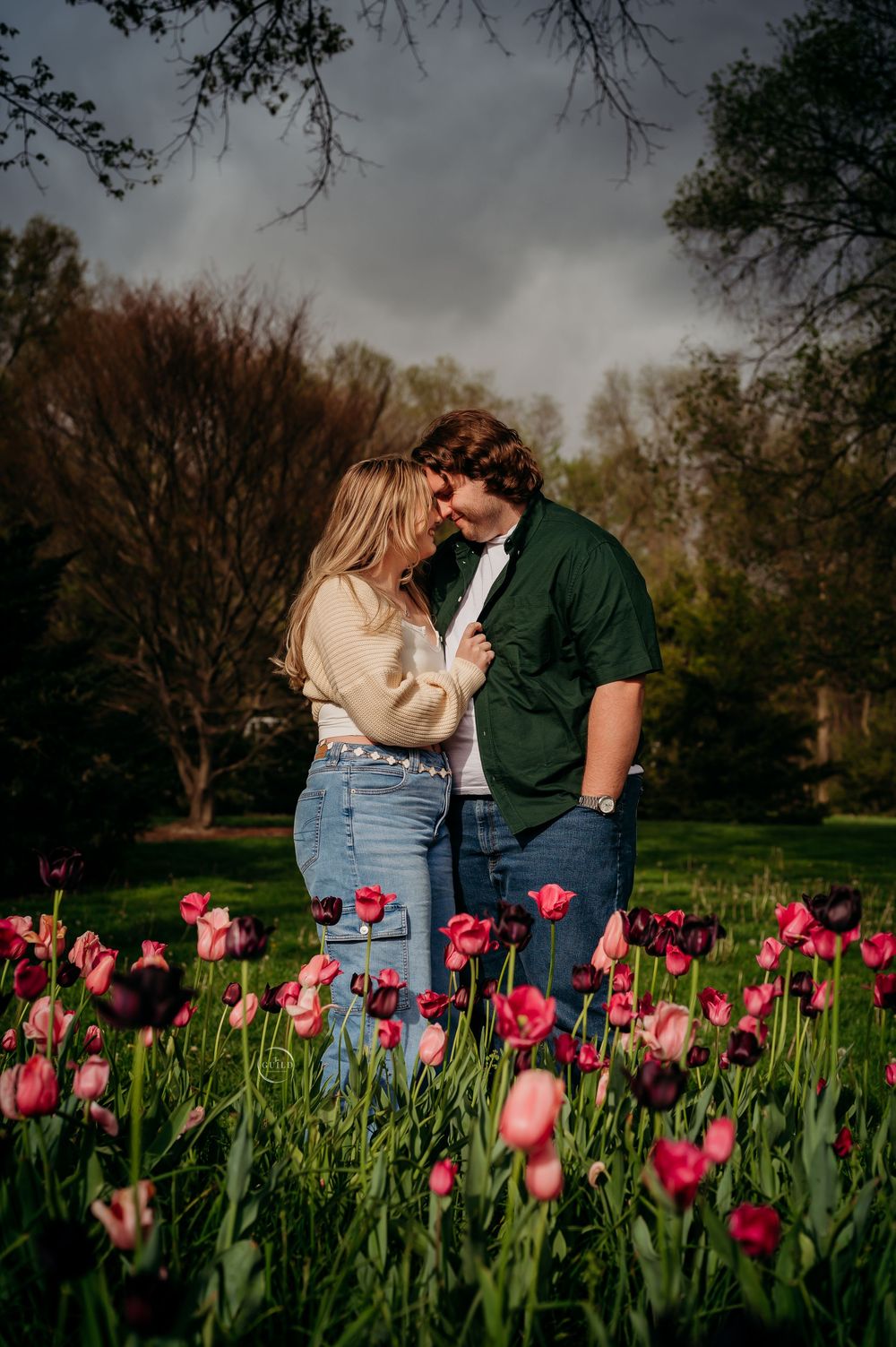 A couple shares a romantic moment surrounded by vibrant pink and red tulips in a lush green park.