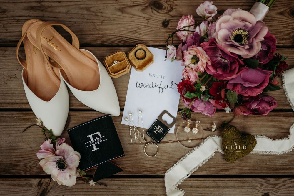 Wedding flat lay with white heels, rings, floral bouquet, and elegant bridal accessories on wood surface.