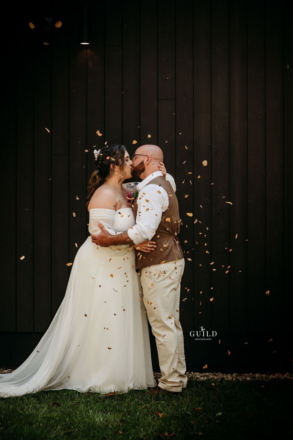 Bride and groom sharing first kiss surrounded by golden confetti against dark barn backdrop.
