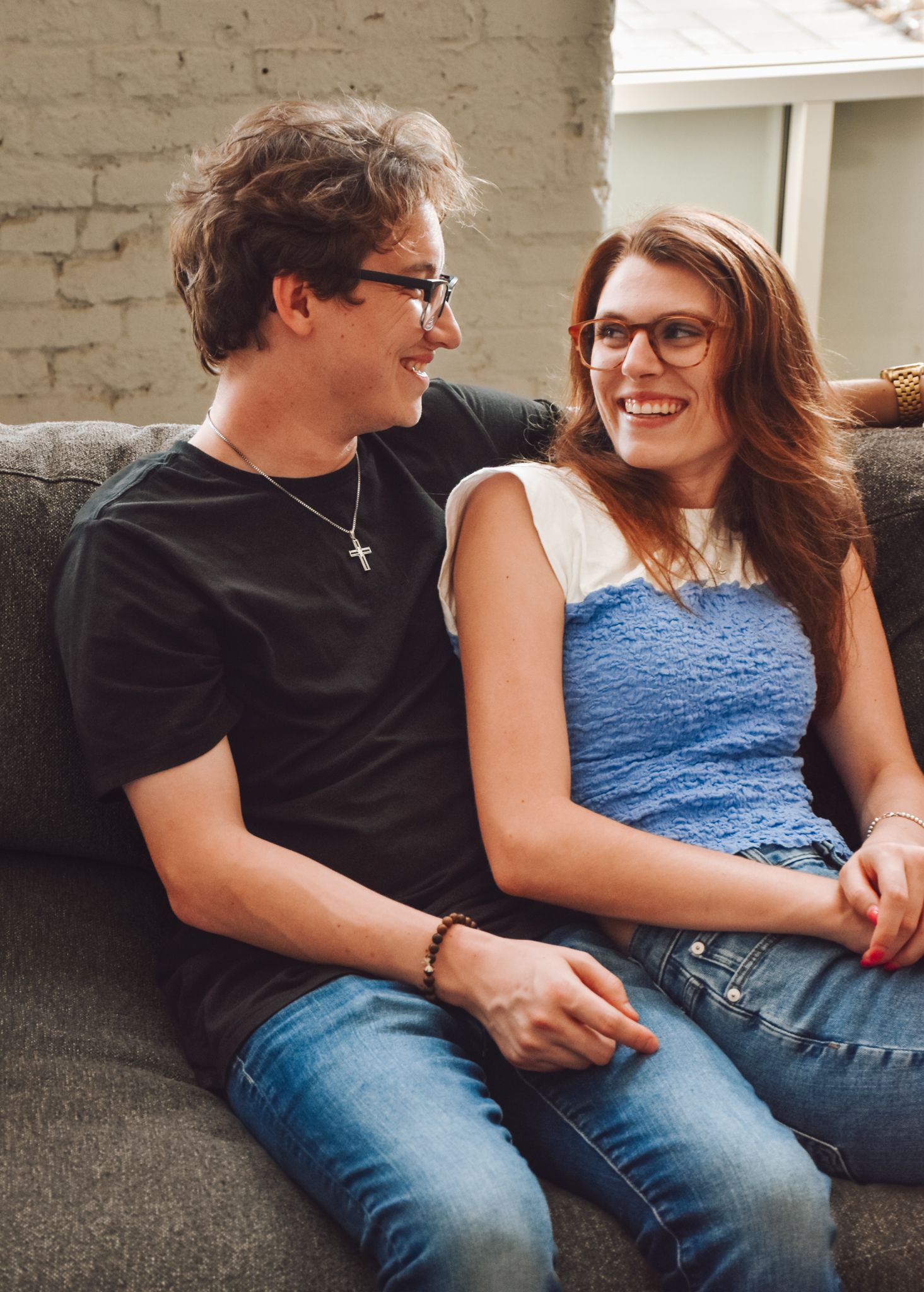 Two people sitting close together on grey couch wearing casual clothes and glasses.