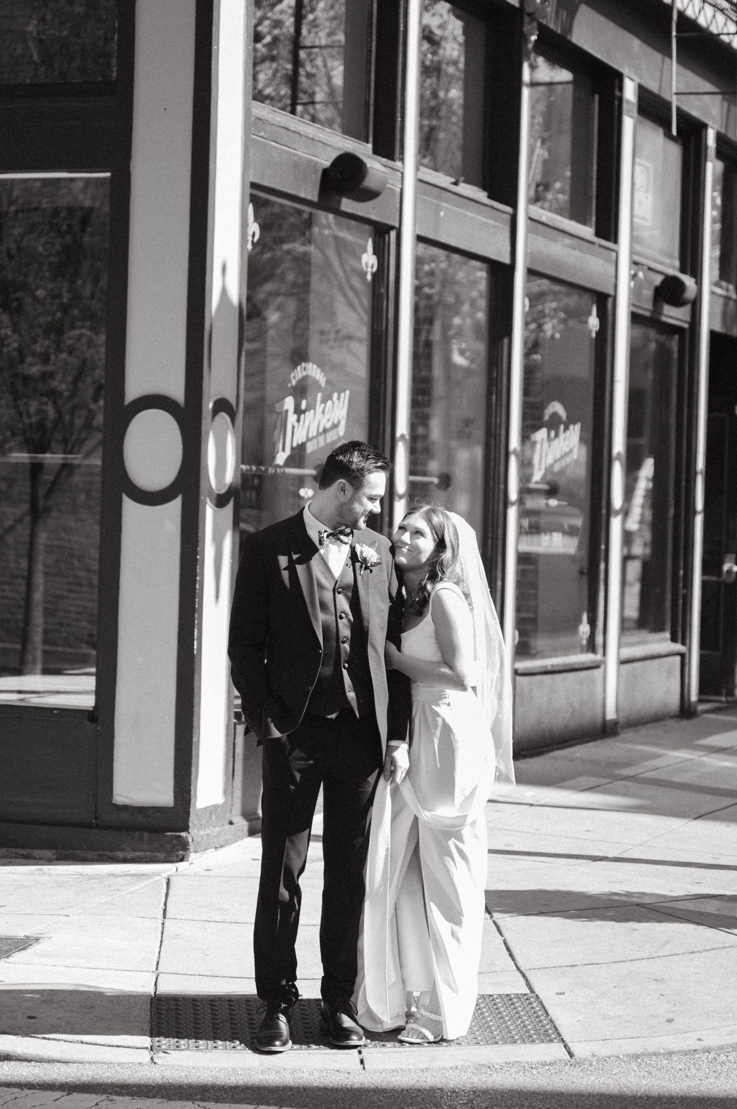 Couple in wedding attire poses together on a city sidewalk in black and white.