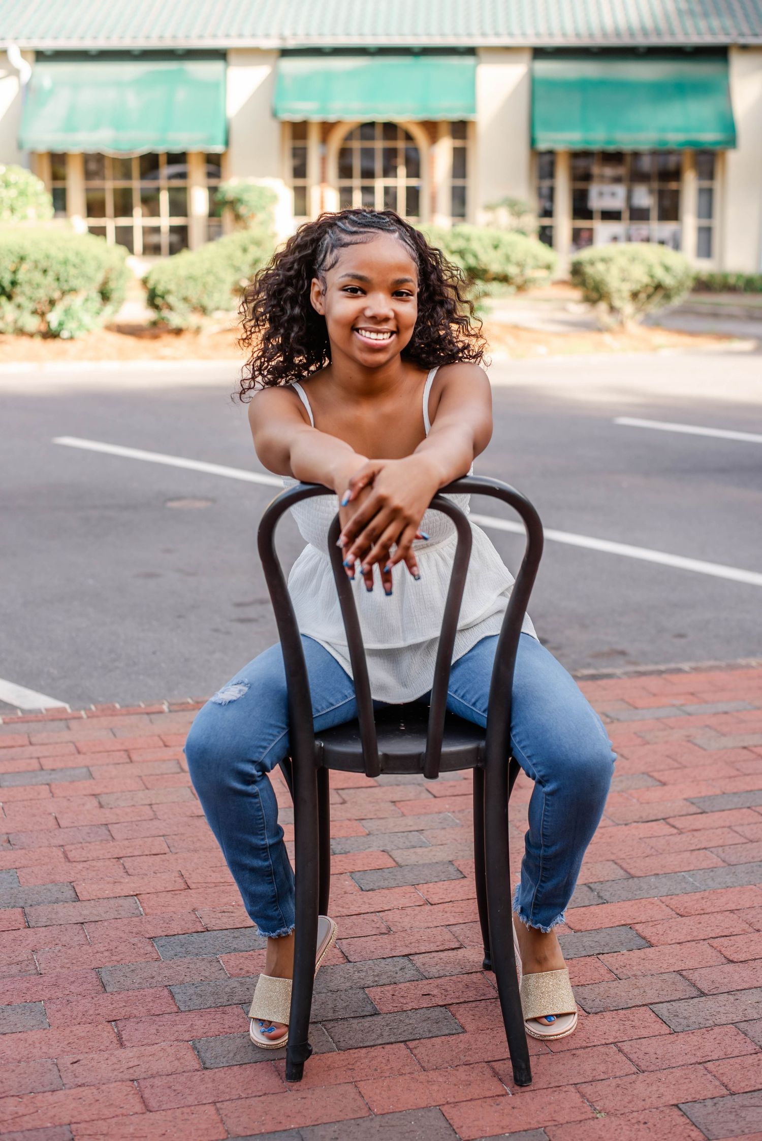 Young woman smiling, sitting backwards on a black chair outdoors on brick pavement, wearing jeans and white top.