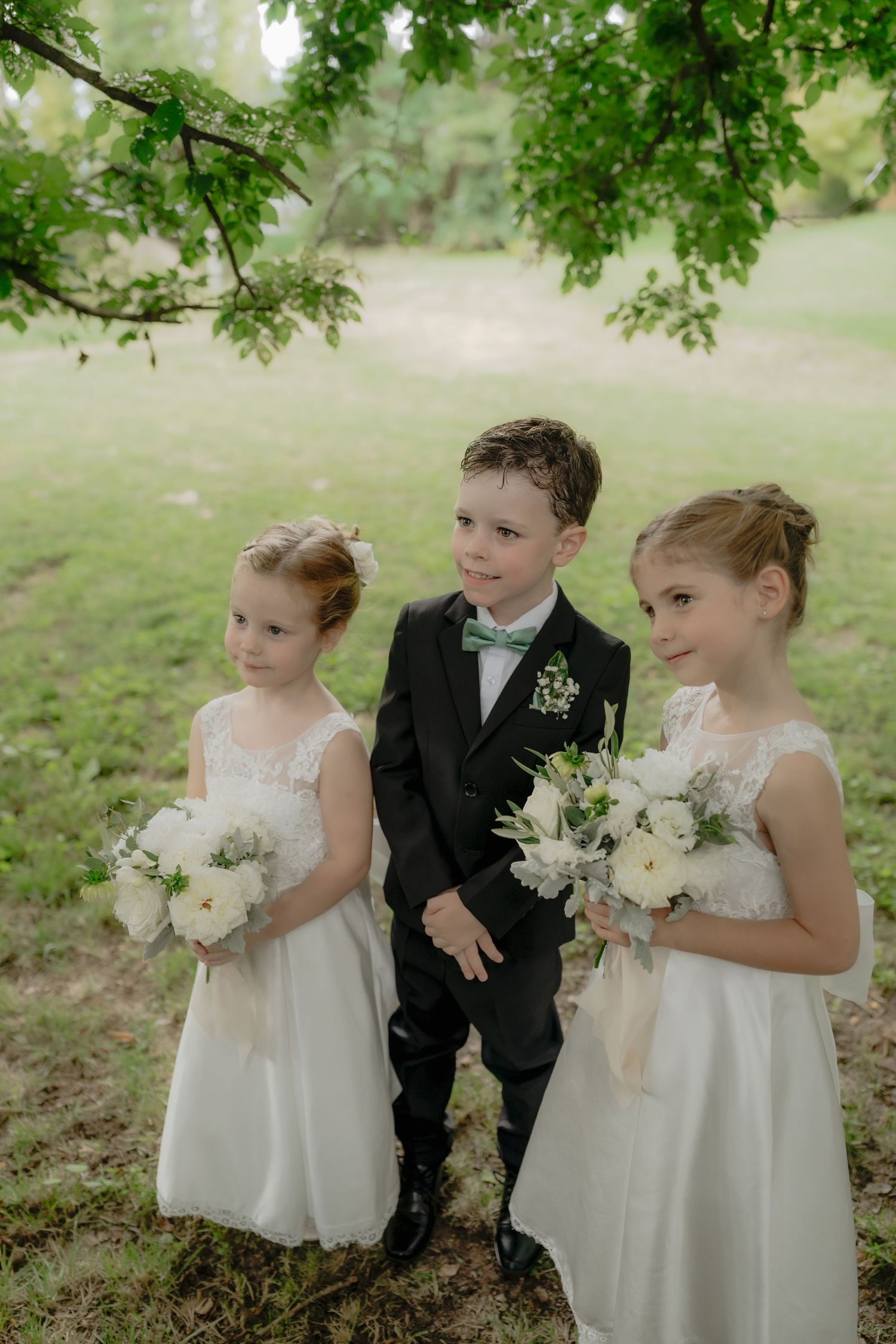 Three children in wedding attire pose outdoors, two flower girls in white dresses holding bouquets flank a ring bearer in a black suit.