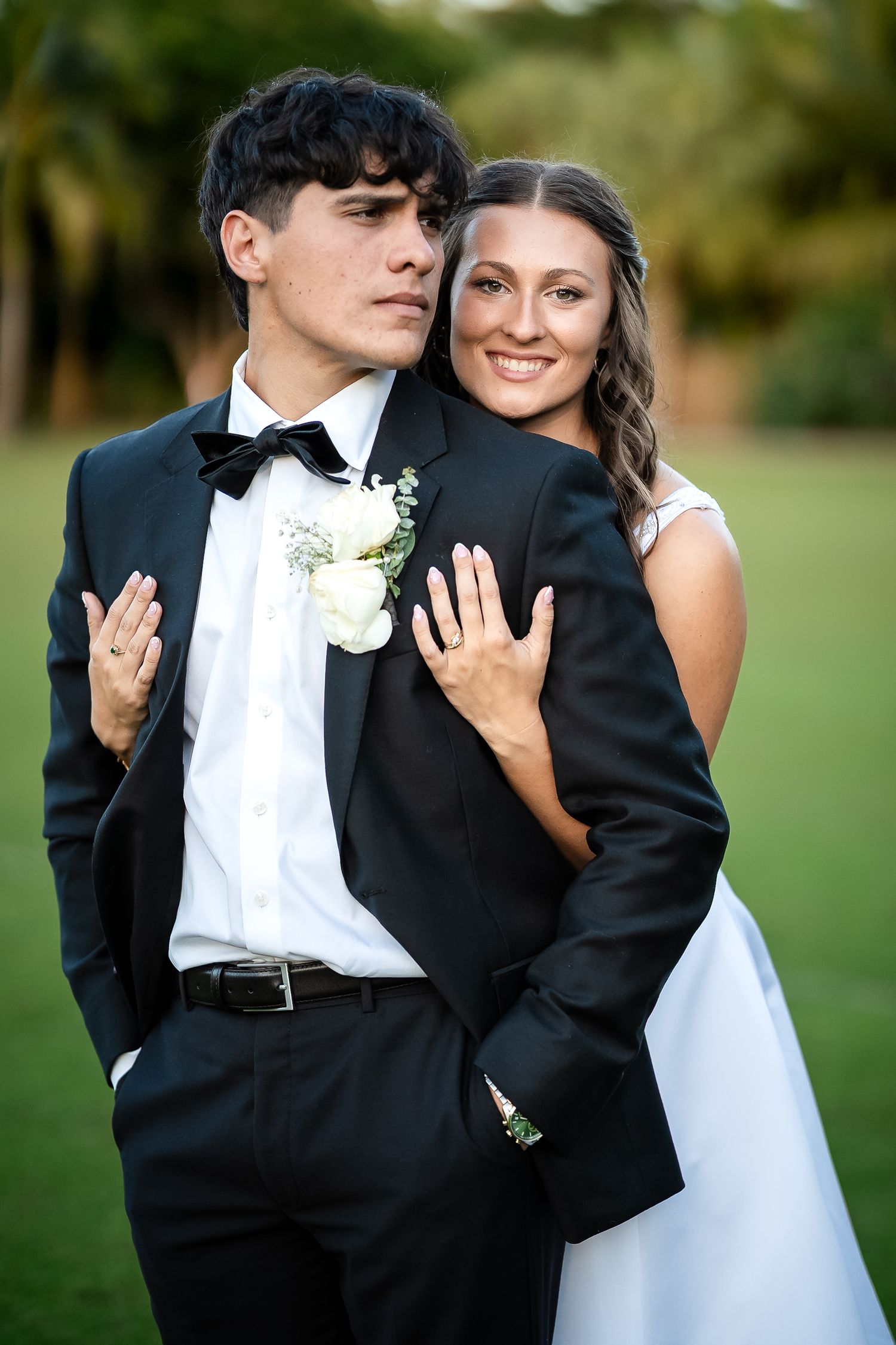 Bride and groom pose outdoors, she embraces him from behind in white dress, he wears black tuxedo with boutonniere.
