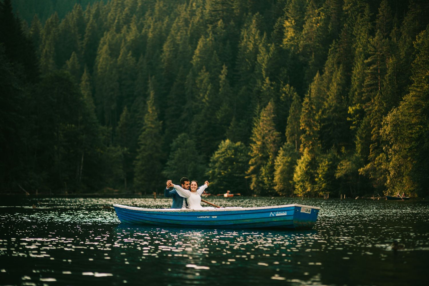 Person joyfully raising arms in a blue canoe on a serene lake surrounded by lush green forest.
