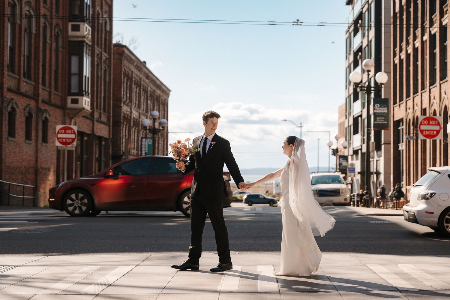 Bride and groom cross a sunny urban street, holding hands, with brick buildings and a red car in the background.