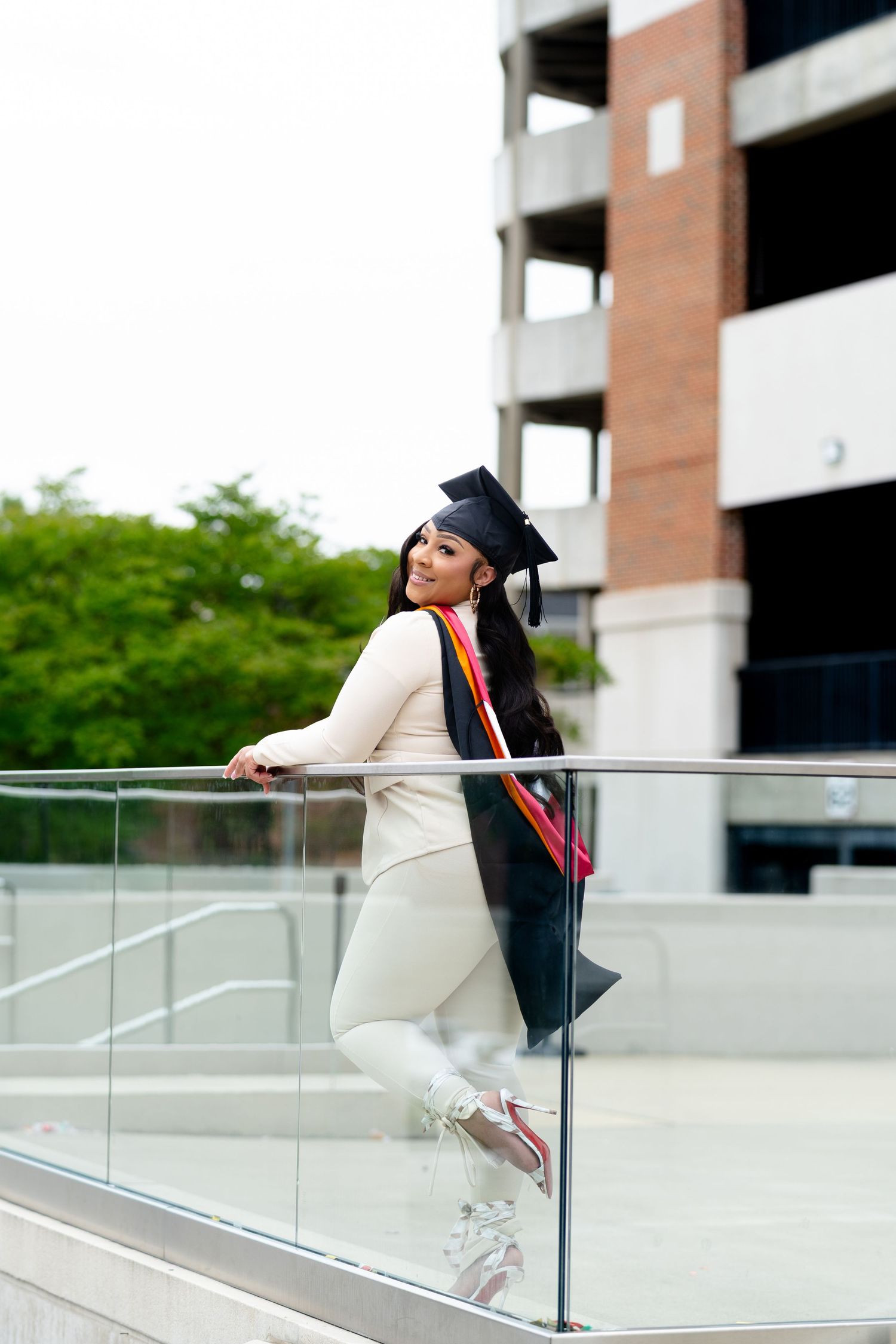 Young woman in graduation cap and gown leans against glass railing outdoors, celebrating academic achievement.