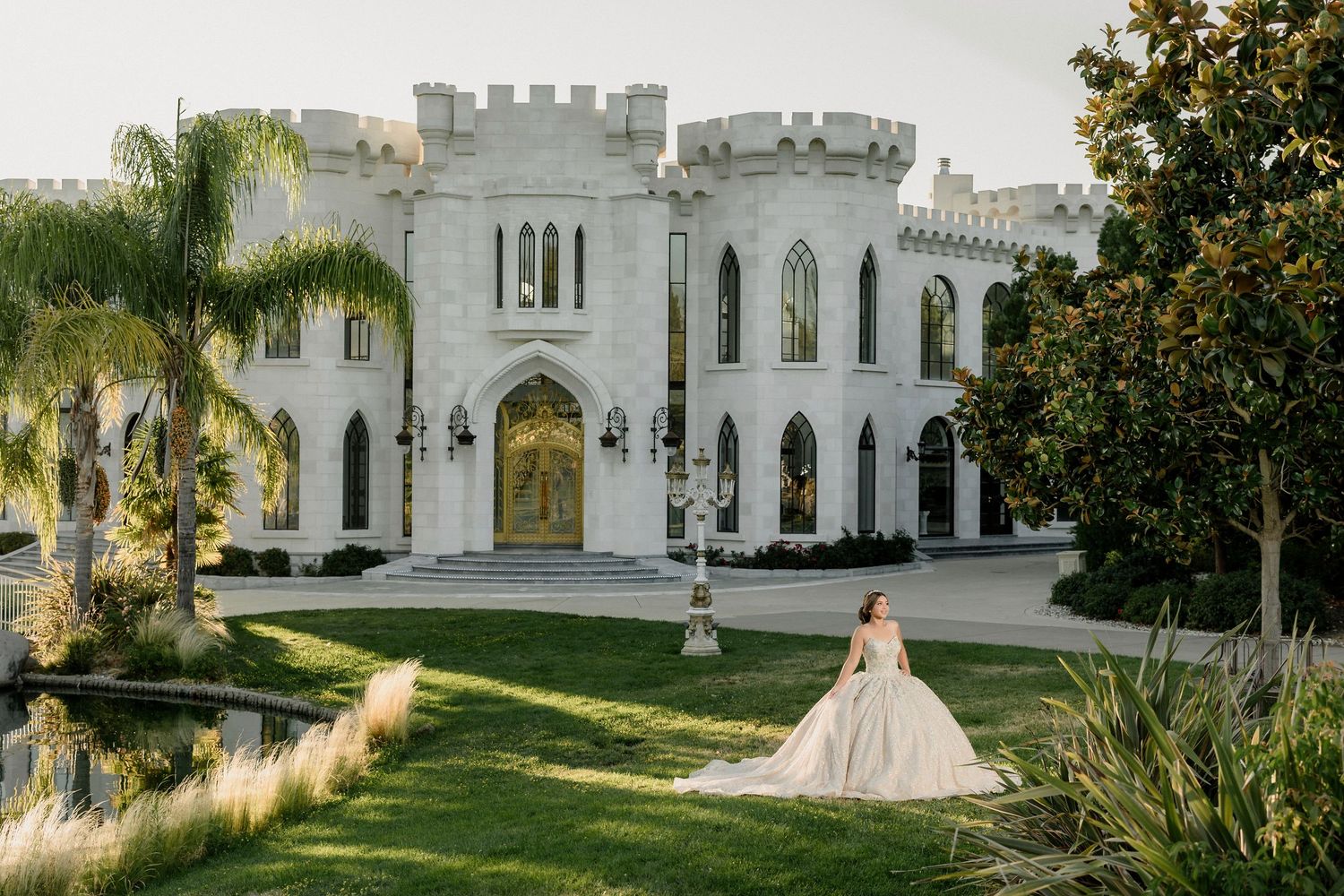 Bride in ballgown poses on lush lawn before a stunning white castle venue with Gothic arched windows and gold doors.