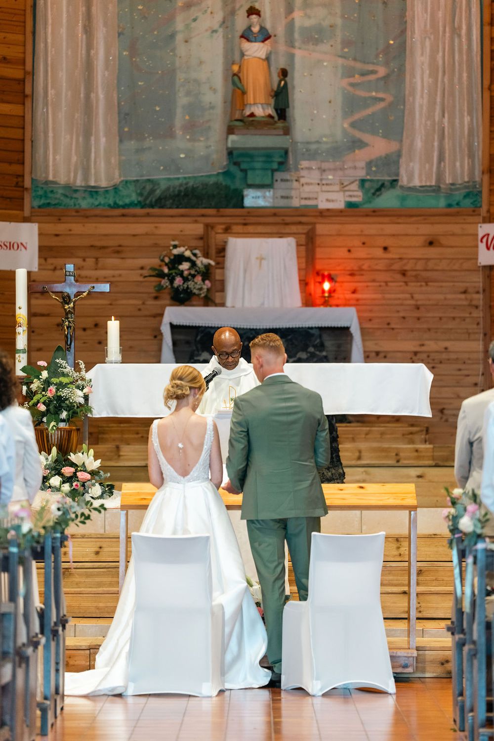 Bride and groom stand at church altar during wedding ceremony with officiant, floral arrangements lining the aisle.